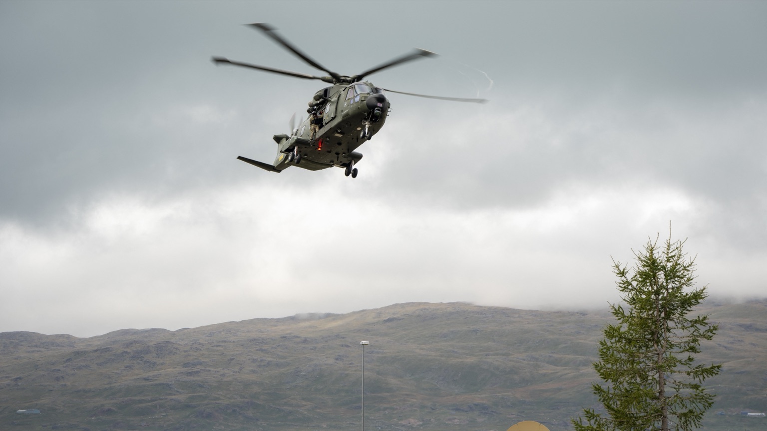 A dark green military transport helicopter in low flight beneath a heavy overcast, rotors blurred, landing gear down. A crew member in camouflage stands in the open side door. Below and behind it lie bare treeless hills and a strip of grey water.