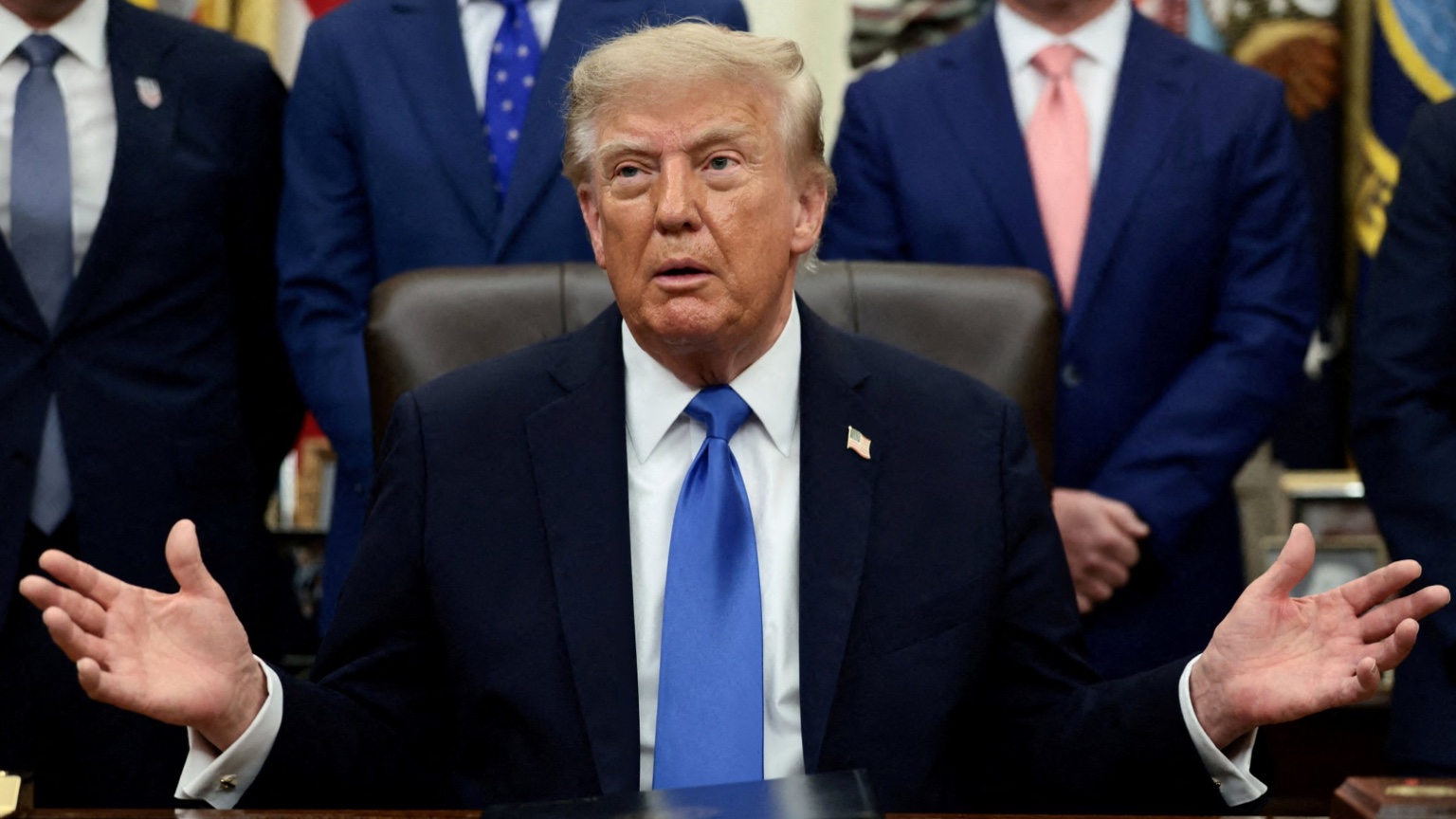 President Trump sits behind the desk in the Oval Office with both hands raised and open in front of him, mid-sentence. He wears a dark navy suit, a white shirt, a blue tie and an American flag lapel pin. Behind him stand several men in dark suits, cropped at the shoulders so their faces are out of frame.